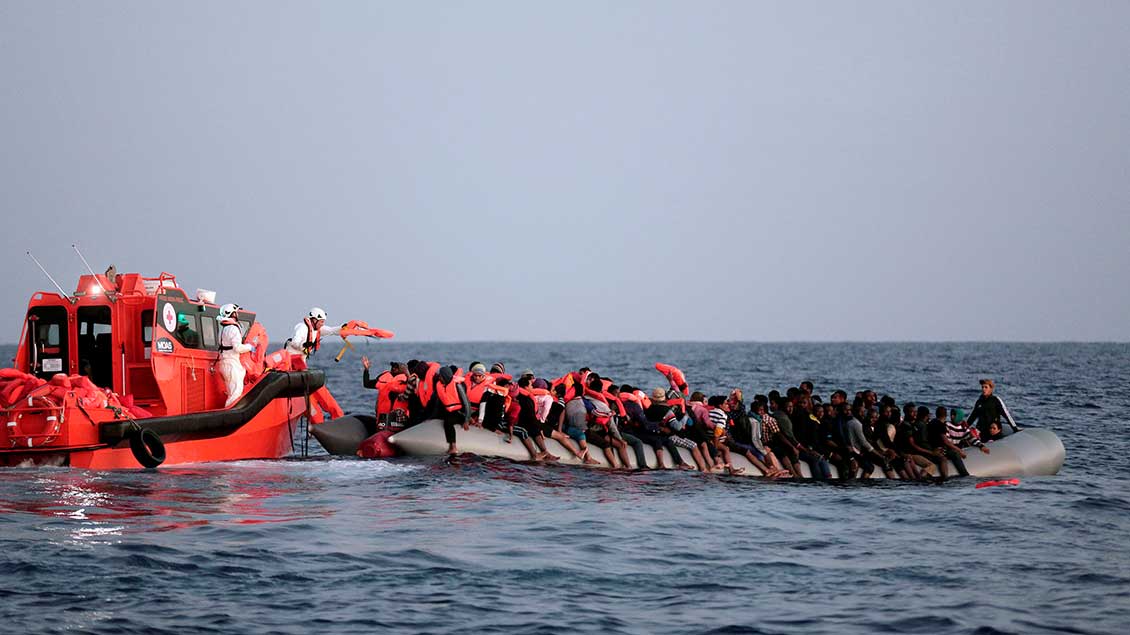 Auf dem Meer schwimmt ein sehr volles Schlauchboot. Daneben ein Rettungsboot. Helfer verteilen Schwimmwesten an die Menschen auf dem Schlauchboot.