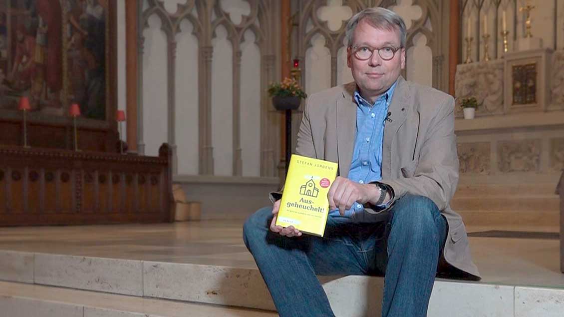 Pfarrer Stefan Jürgens auf den Stufen im Altarraum der Heilig-Kreuz-Kirche in Münster. In den Händen hält er sein neues Buch "Ausgeheuchelt".