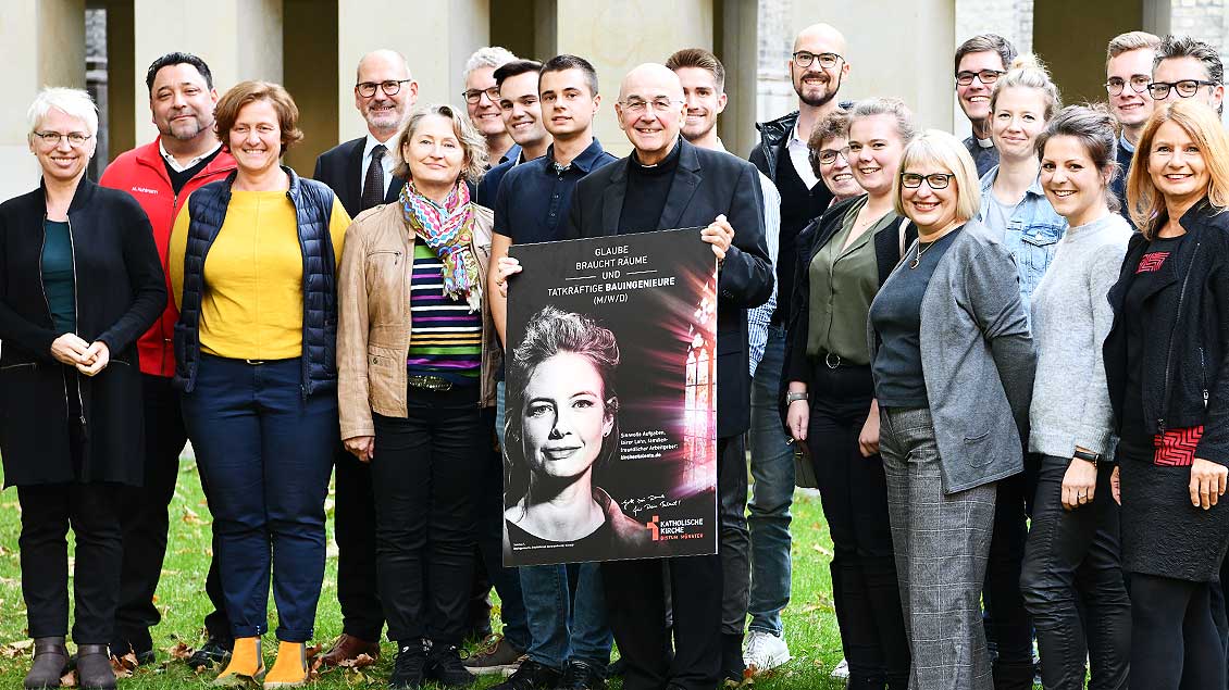 Bischof Felix Genn steht mit einem Plakat der Arbeitgeber-Kampagne unter vielen Mitarbeiter, die als Foto-Models bei der Kampagne mitgemacht haben.