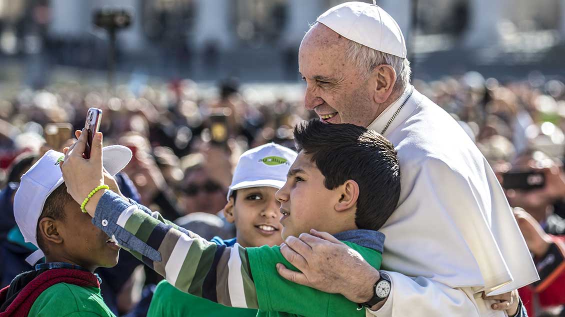 Drei Jungen machen mit einem Smartphone ein Foto mit Papst Franziskus auf dem Petersplatz.