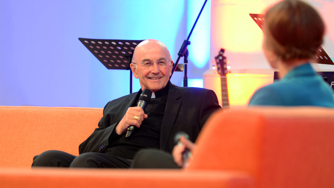 Bischof Felix Genn sitzt in der Jugendkirche in Münster auf einem Sofa und spricht in ein Mikrofon.
