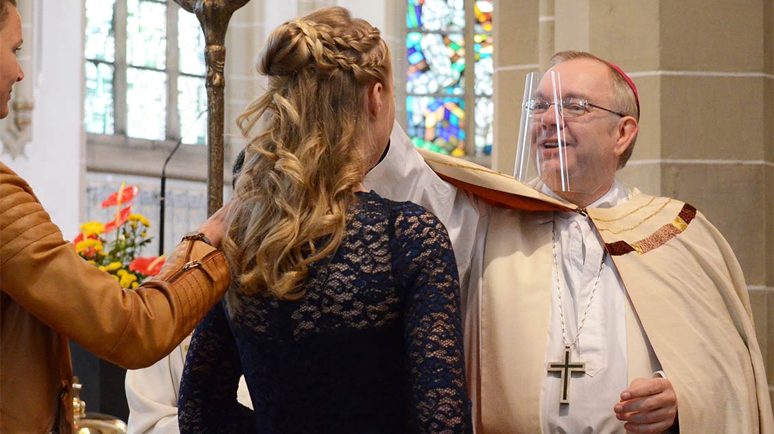 Weihbischof Christoph Hegge trägt einen Gesichtsschutz und spendet einer jungen Frau den Segen.