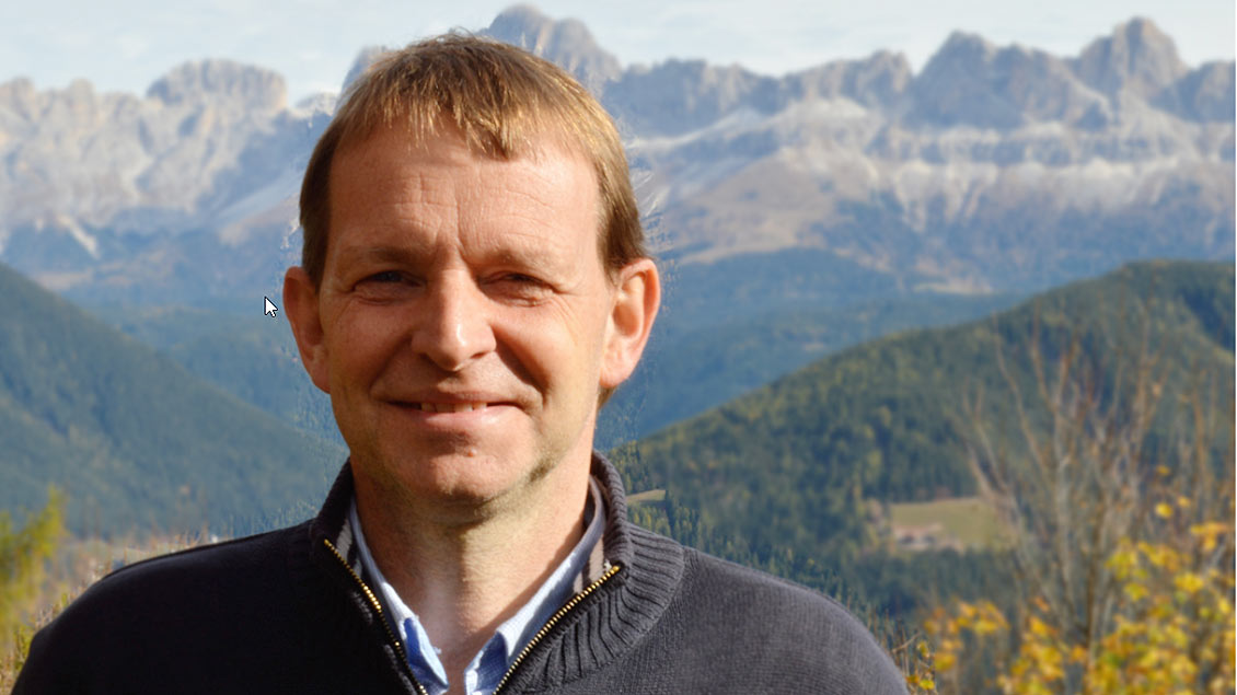 Andreas Rehm steht vor einem Bergpanorama und lächelt.