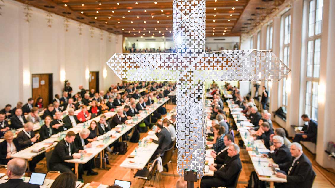 Blick durch ein gelochtes Metallkreuz auf die Teilnehmer an langen Tischen bei den Beratungen der Synodalversammlung am 31. Januar 2020 im Dominikanerkloster in Frankfurt.