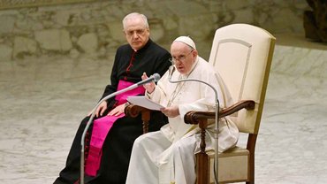 Papst Franziskus spricht in zwei Mikrofone. Rechts neben ihm sitzt eine schwarz gekleidete Person.