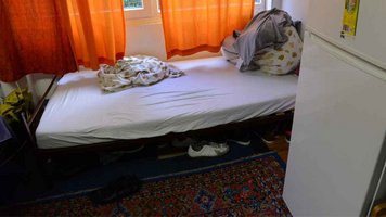 Ein unaufgeräumtes Bett. Orangene Gardinen hängen vor dem Fenster. Unter dem Bett stehen Schuhe. 