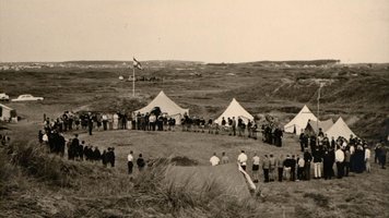 Ein Schwarz-/Weißbild. Viele Menschen stehen in einem Kreis in den Dünen. Es sind Zelte zu sehen.