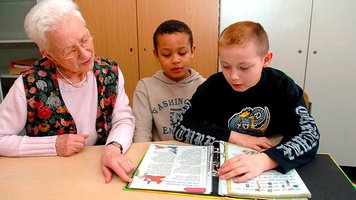 Eine ältere Frau unterstützt zwei Schulkinder beim Lesen.