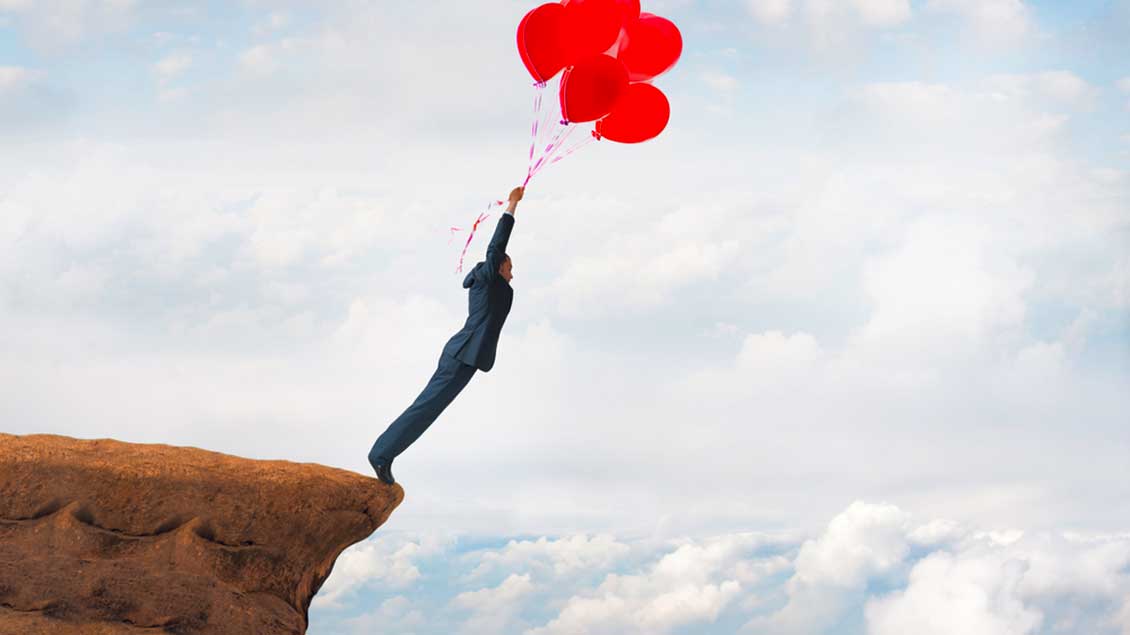 Ein Mann steht mit Luftballons in der Hand am Rand einer Klippe.