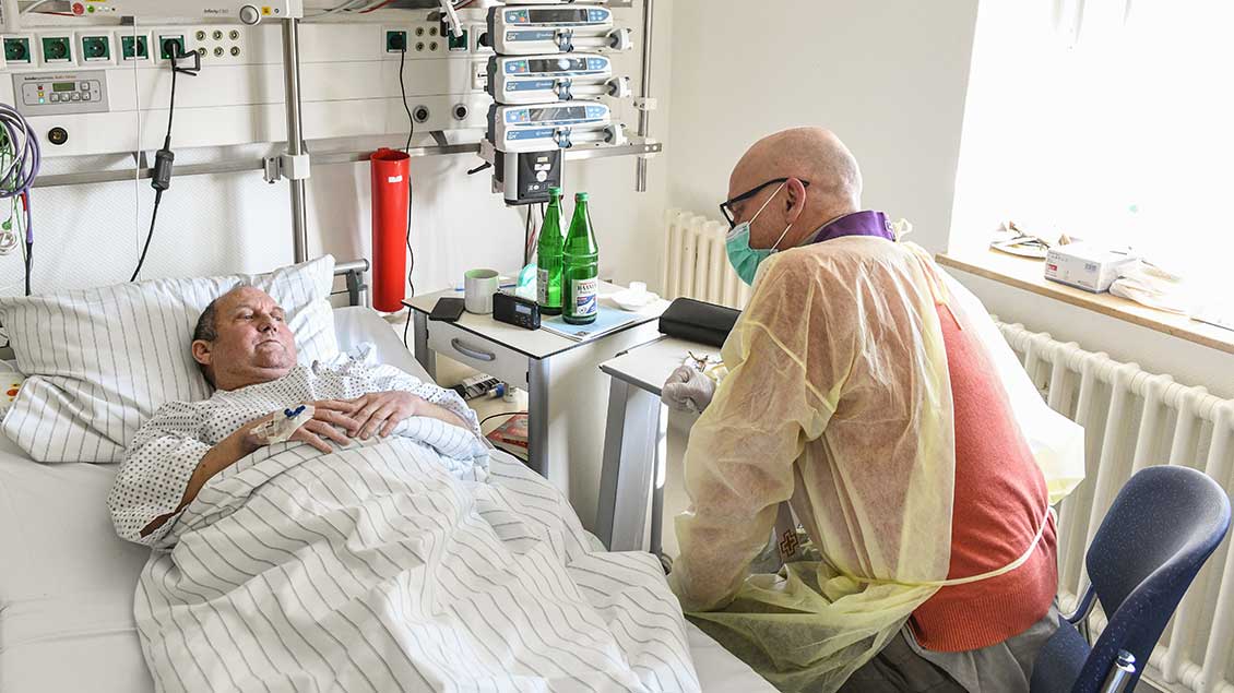 Ein Klinikseelsorger mit Schutzkittel, Atemschutzmaske und Gummihandschuhen und einer Stola spricht mit einem Patienten im Krankenbett bei der Durchführung der Krankenkommunion