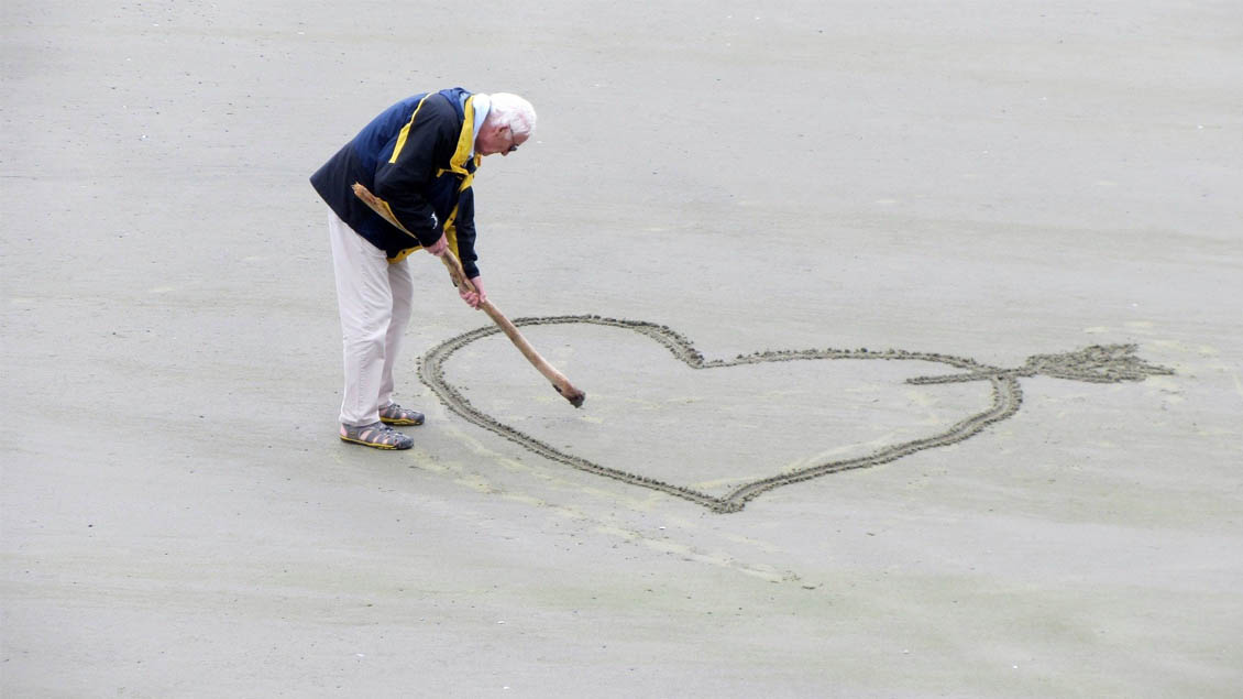 Älterer Mann zeichnet ein Herz in den Sand.