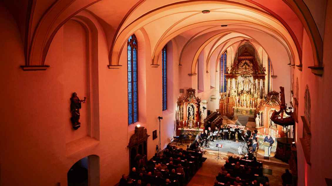 Eine Kirche von innen. Die Wände sind rotlich angestrahlt. Am Altar steht ein Orchester. Die Bänke sind mit Menschen gefüllt.