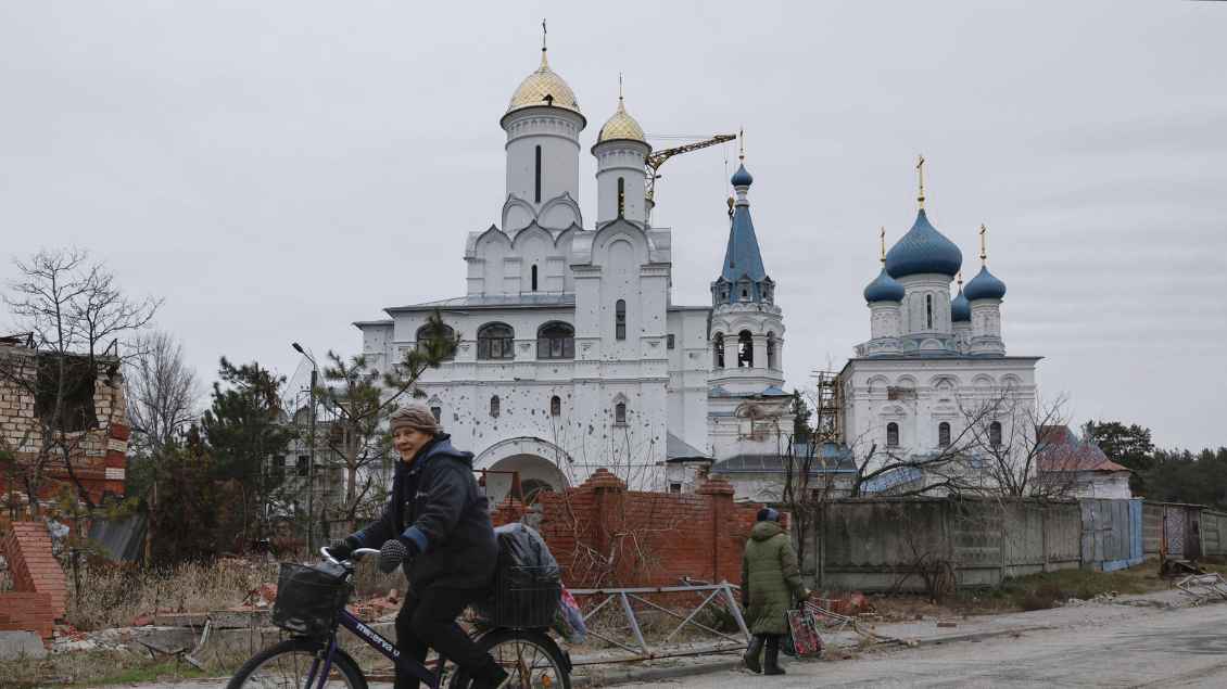 Eine orthodoxe Kirche, in dessen weißer Fassade viele Einschusslöcher zu sehen sind. Vor der Kirche fährt eine Frau auf einem Fahrrad. Daneben geht eine Frau zu Fuß.