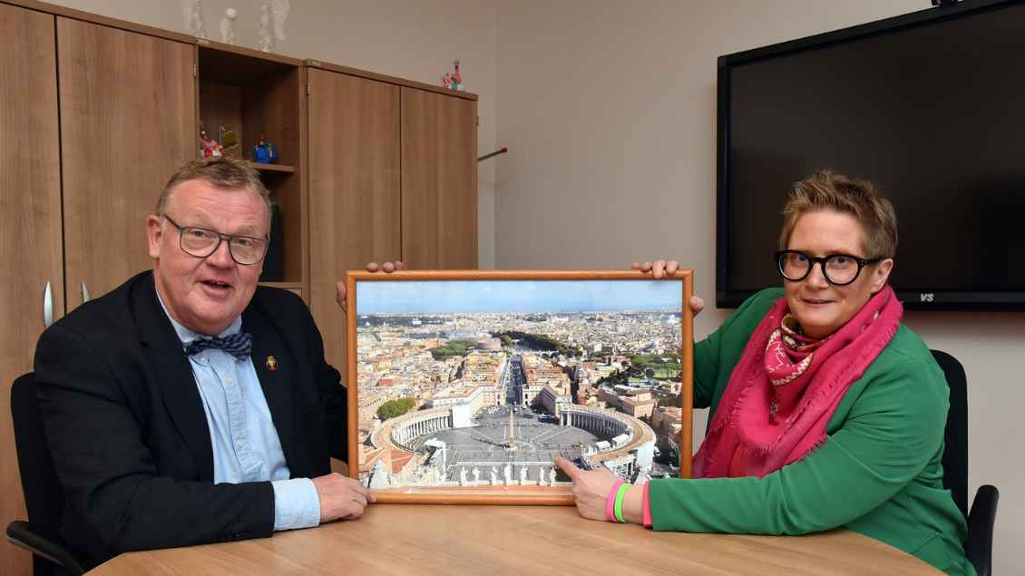 Zwei Personen an einem Tisch. Sie halten in der Mitte eine Bild vom Petersplatz in die Höhe. Foto: Ludger Heuer (Schulstiftung St. Benedikt)