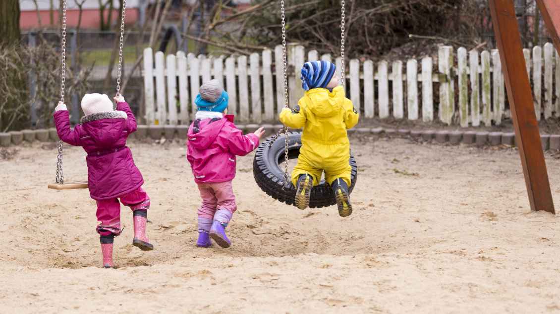 Drei Kinder spielen im Sand an einer Schaukel.
