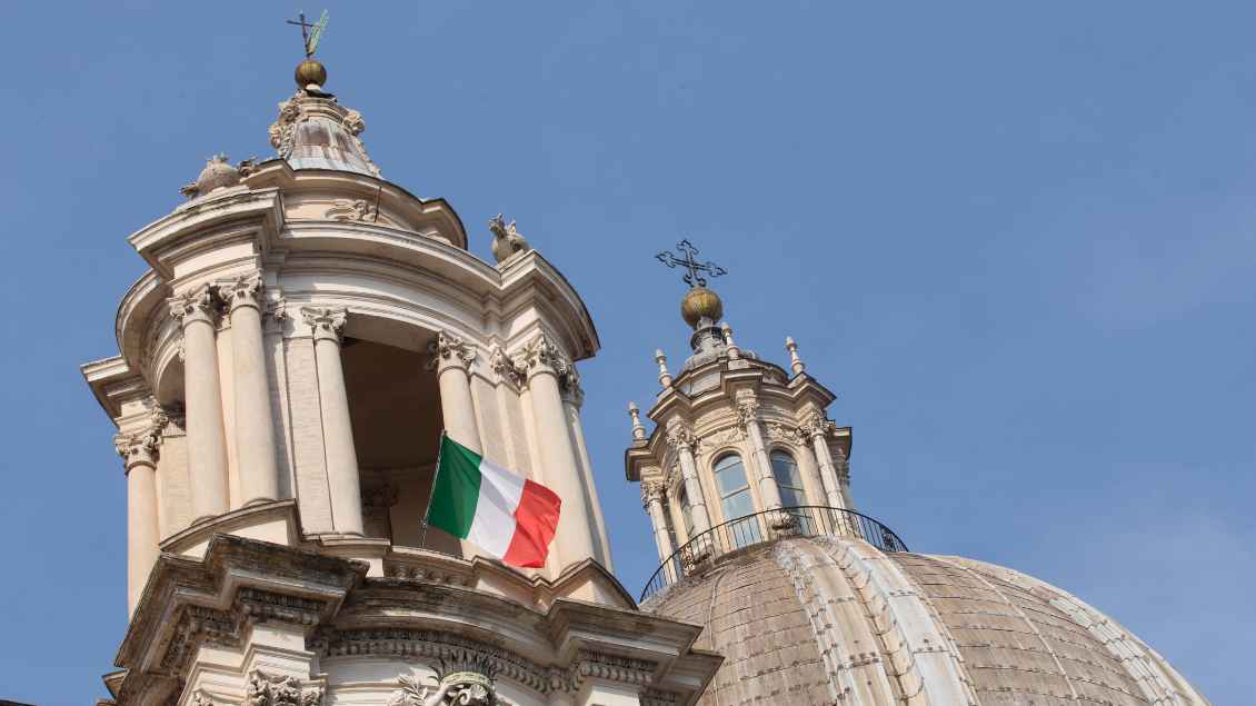 Zwei Türme einer Kirche aus Stein. Eine italienische Flagge weht aus dem Fenster. Blauer Himmel hängt über der Kirche.