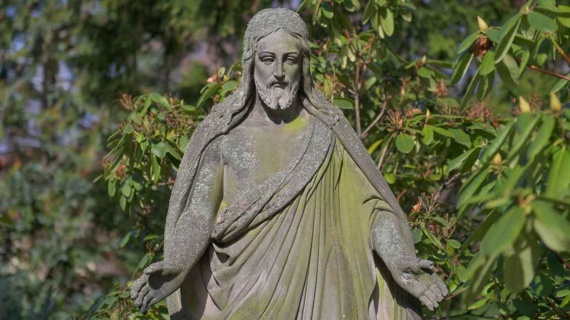 Eine Jesus-Statue in der Natur aus Stein. Die Statue ist leicht mit Moos bedeckt.