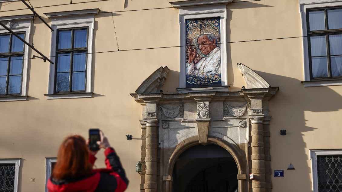 Eine Frau in roter Jacke fotografiert mit ihrem Handy ein Gemälde von Papst Joahnnes Paul II. Das Gemalde hängt über einem Torbogen einer gelben Hauswand.