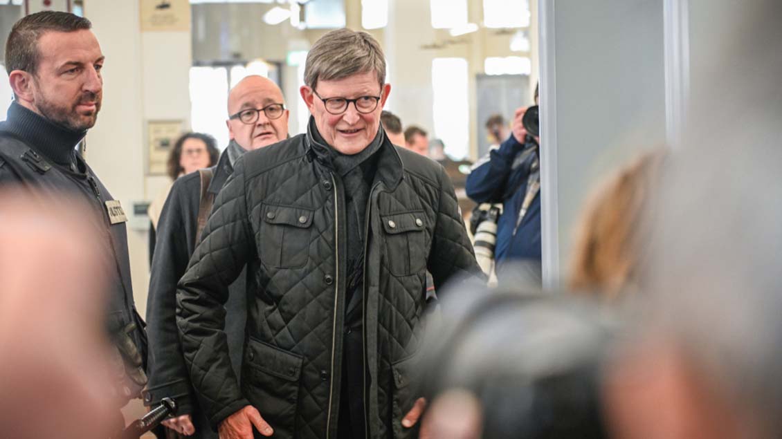 Kardinal Woelki steht neben einem Mitarbeiter der Justiz an einer Sicherheitskontrolle am Kölner Landgericht. Woelki lächelt und hat eine schwarze Jacke an. Im Vorder- und Hintergund sind Fotografen zu sehen.