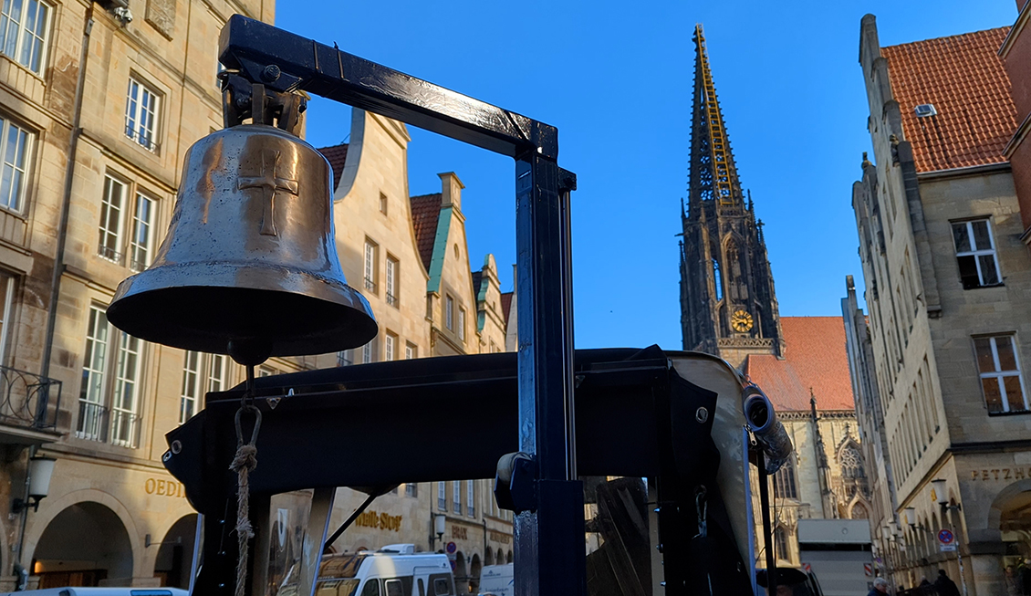 Eine Glocke auf dem Wagen. Im Hintergrund ist die Stadtkulisse Münsters mit der St.-Lamberti-Kirche.