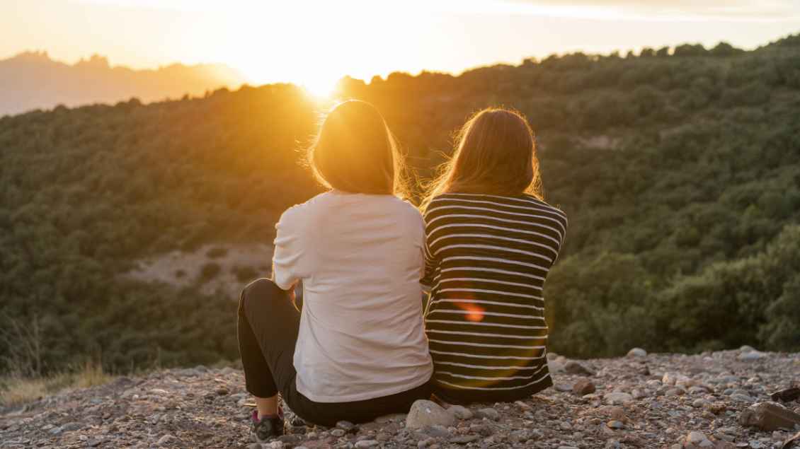 Zwei junge Frau sitzen auf einem Felsen und blicken auf einen Sonnenuntergang Foto: Westend61 (Imago)