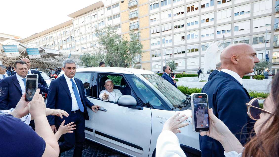 Papst Franiskus sitzt in einem weißen Auto und winkt aus dem offenen Fenster. Um das Auto herum stehen Bodyguards und viele Schaulustige.