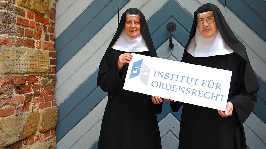 Schwester Lydia und Schwester Scholastika mit einem Schild "Institut für Ordensrecht" vor dem Kloster Dinklage