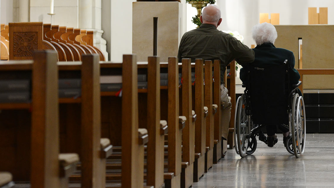 Rollstuhlfahrer steht im Mittelgang einer Kirche, daneben sitzt ein Mann in der Bank