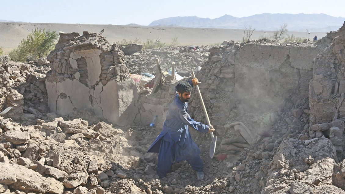Ein Afghane in blauem Mantel steht in den Trümmern eines Gebäudes mit einer Schaufel.