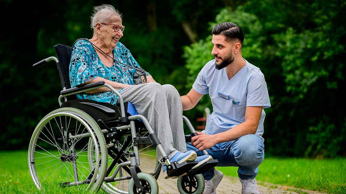 Pfleger unterhält sich knieend mit einer Frau im Rollstuhl