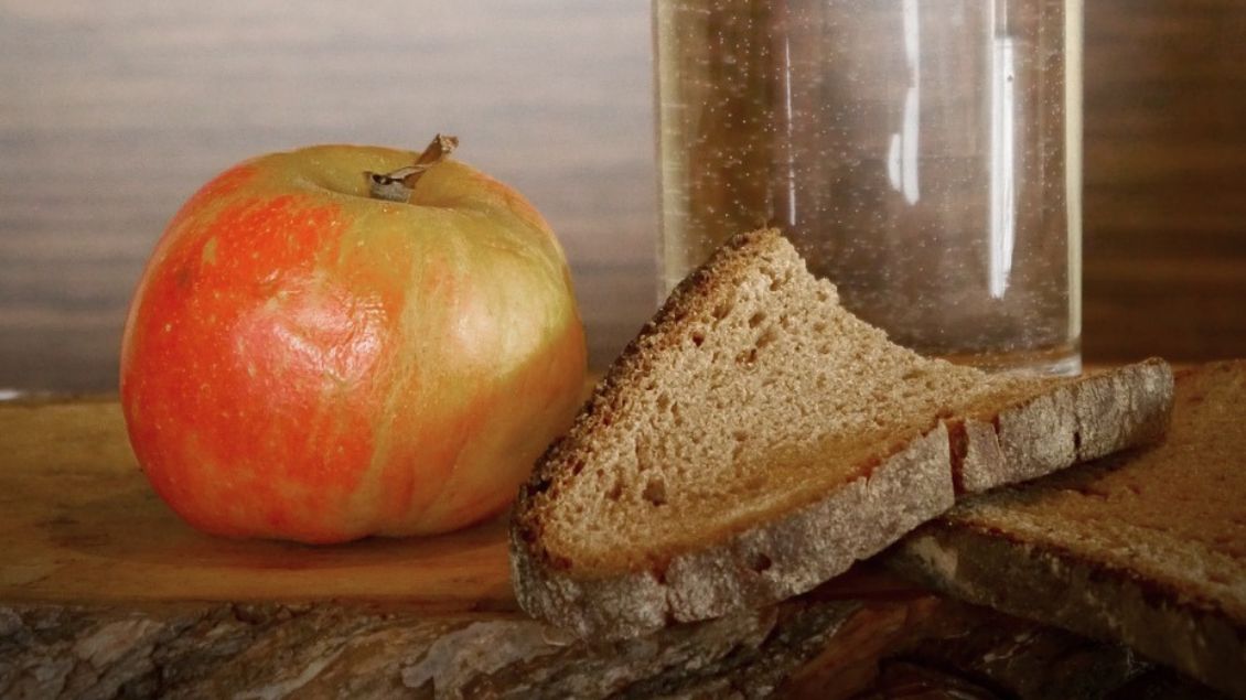 Ein Apfel liegt neben einer trockenen Scheibe Brot. Dahinter steht ein Glas Wasser.